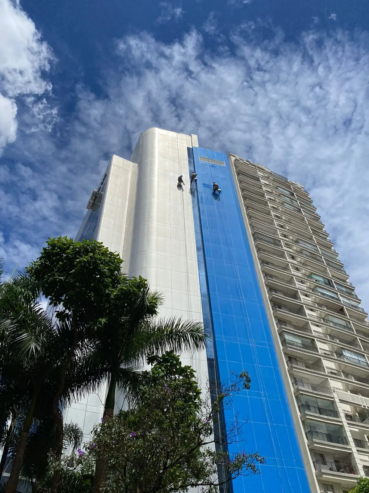 Equipe técnica realizando pintura de fachada predial suspensa por cordas